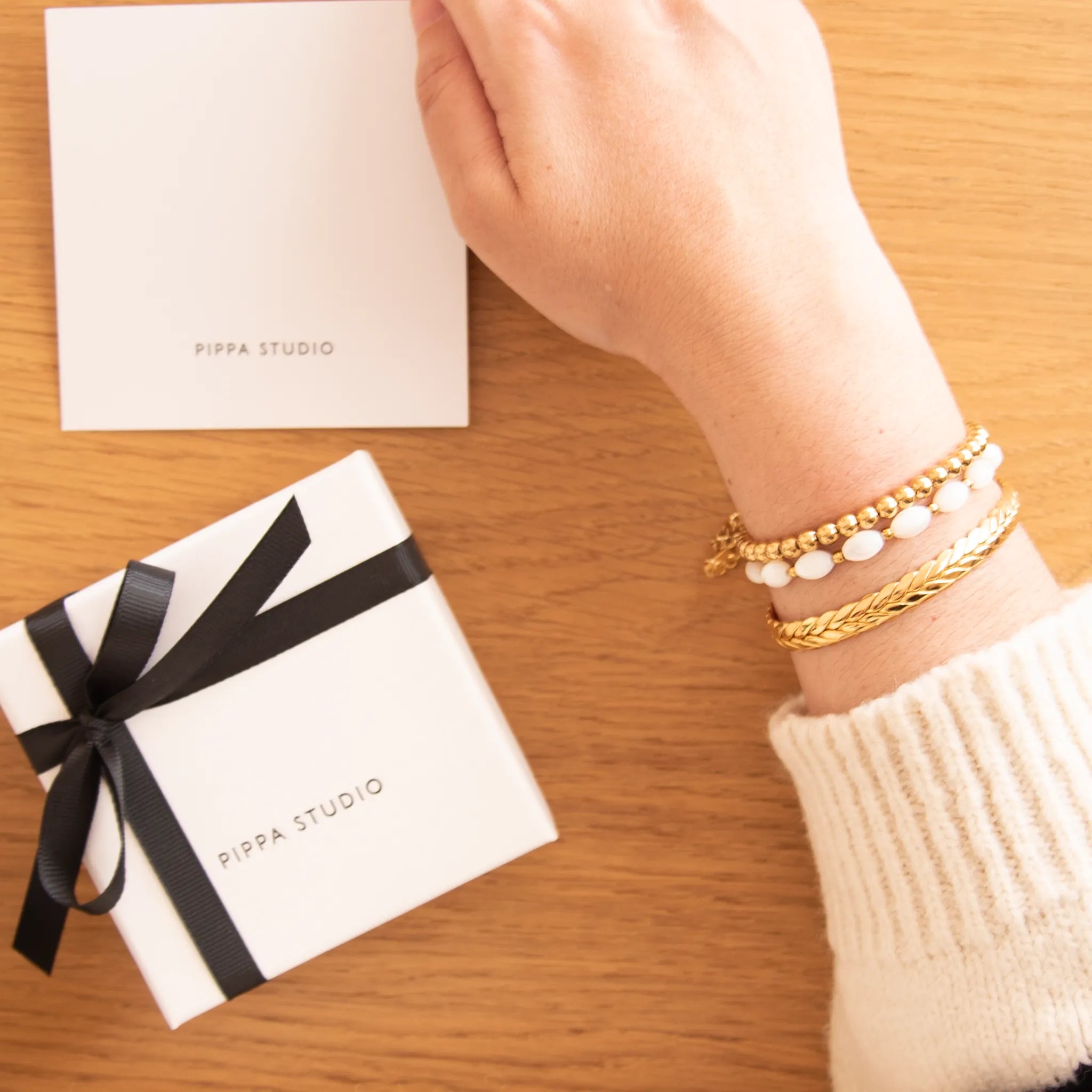 Hand wearing gold bracelets with a Pippa Studio gift box on a wooden surface