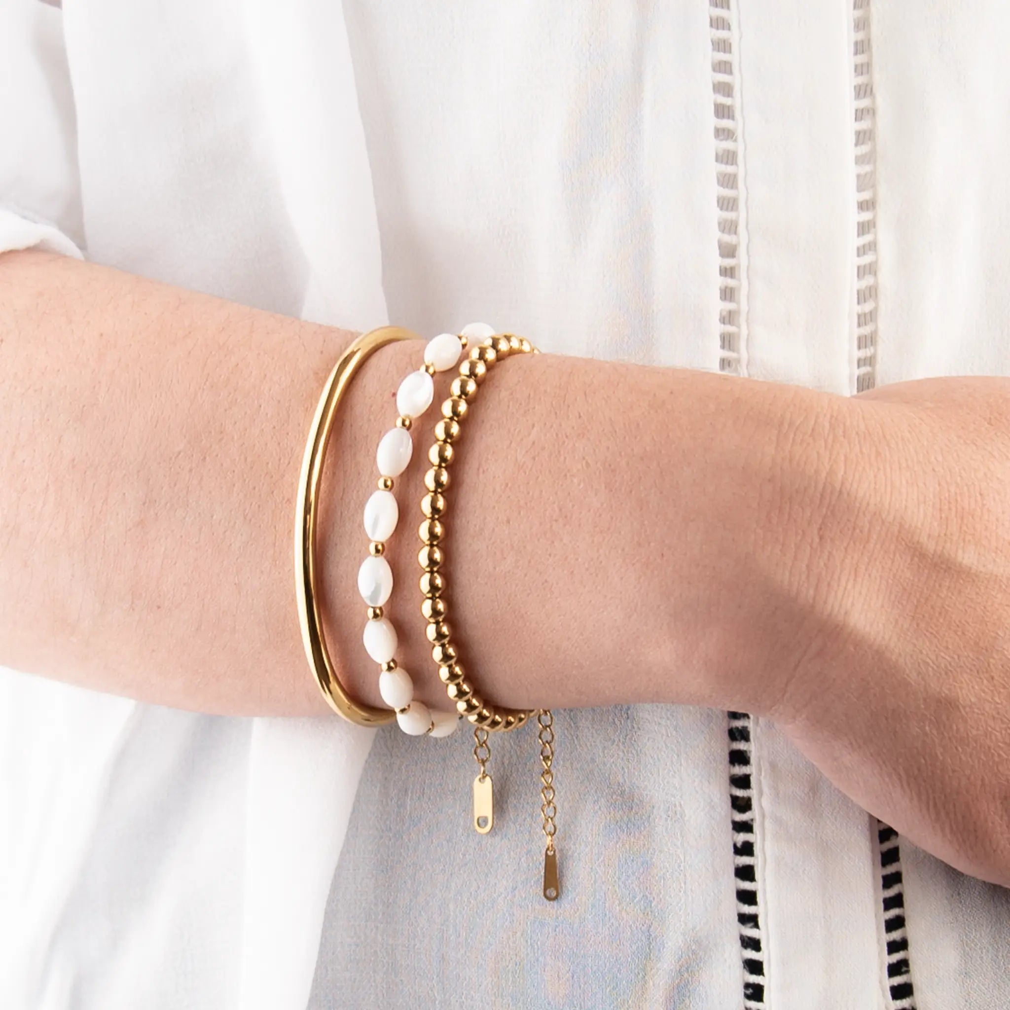 Three golden bracelets, one with white beads, on a person with a white shirt