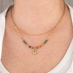 Golden necklace with green and red beads and a sun pendant on a person wearing a white shirt.