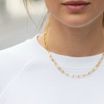 Gold paperclip chain necklace worn by a person against a neutral background