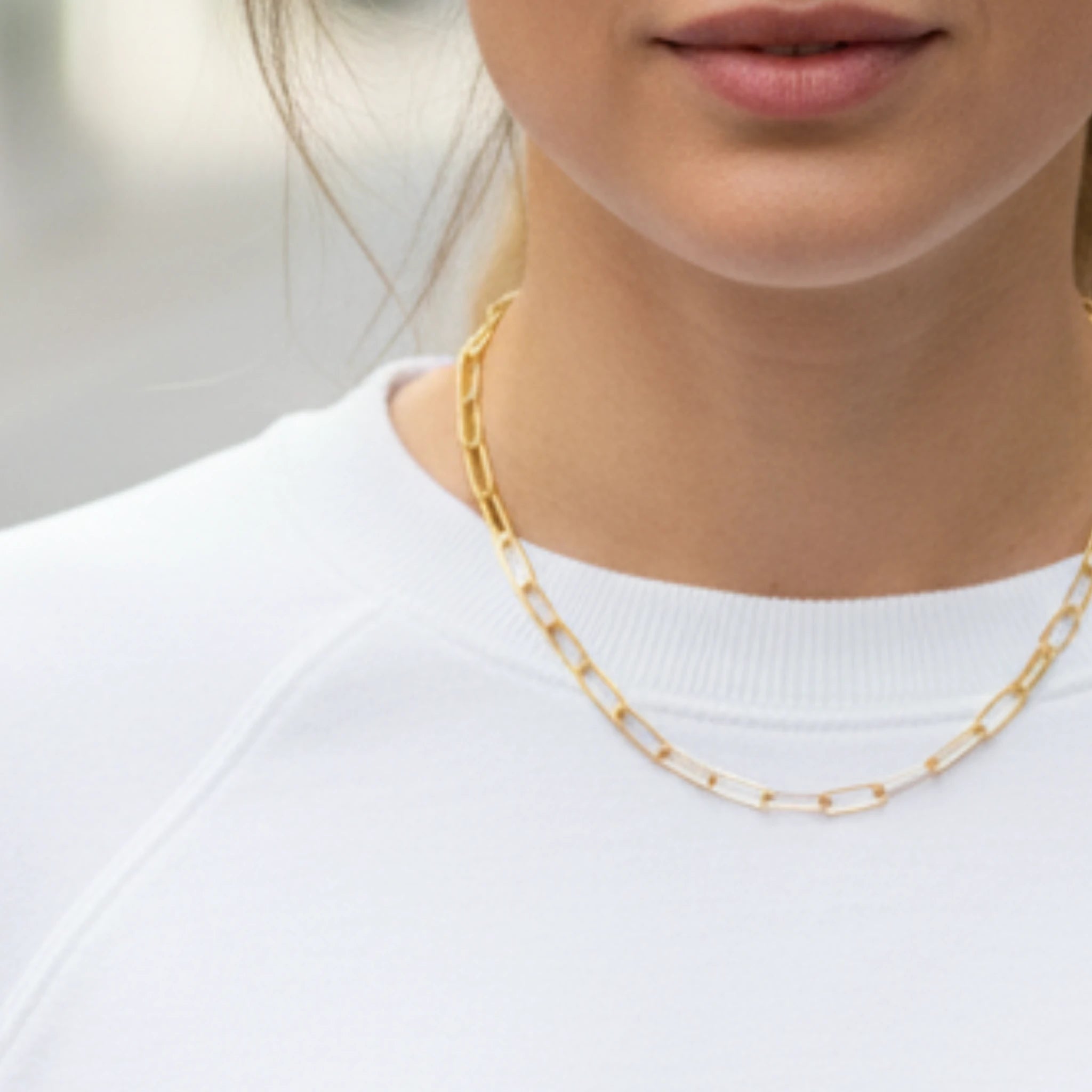 Gold paperclip chain necklace worn by a person against a neutral background