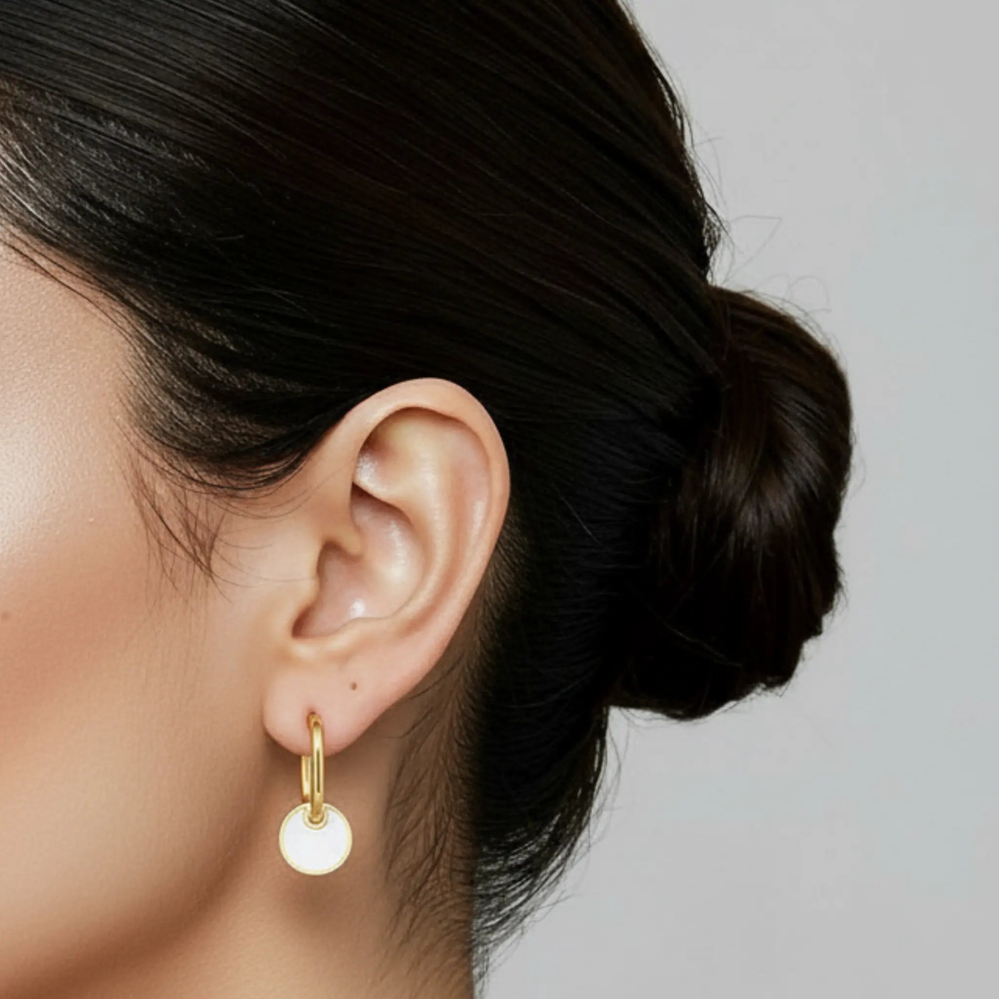 Close-up of an ear wearing a gold hoop earring with a white round charm against a neutral background