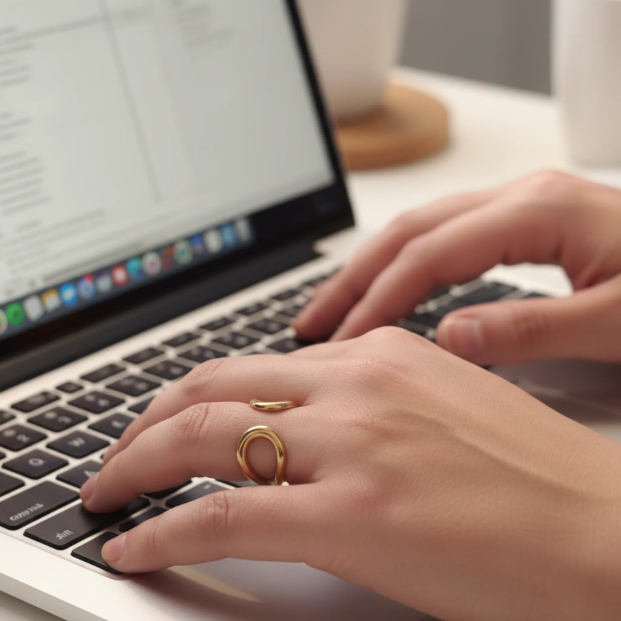 Close-up of hands typing on a laptop with a gold open front sculptural ring on a blurred background